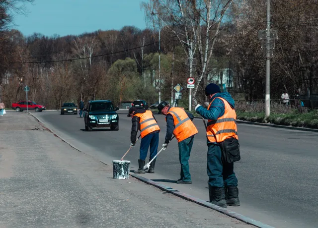 Three road workers in reflective vests paint street markings on a suburban road. Parking lot striping staff costs explained