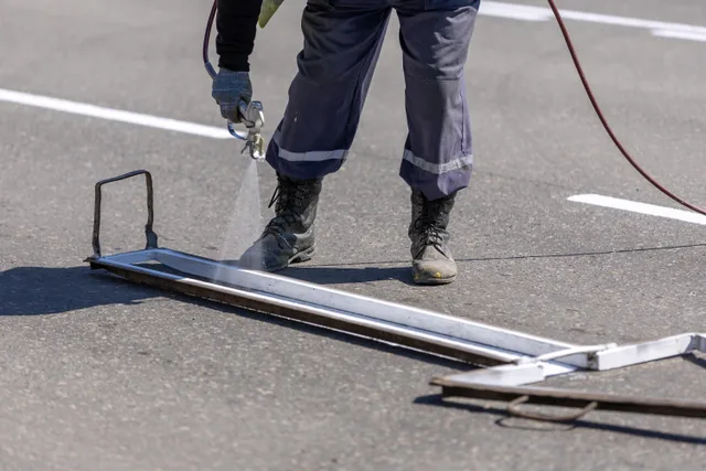 A construction worker painting white lines on a road with a spray gun during the day. Parking lot striping prices and margins for owners in 2027